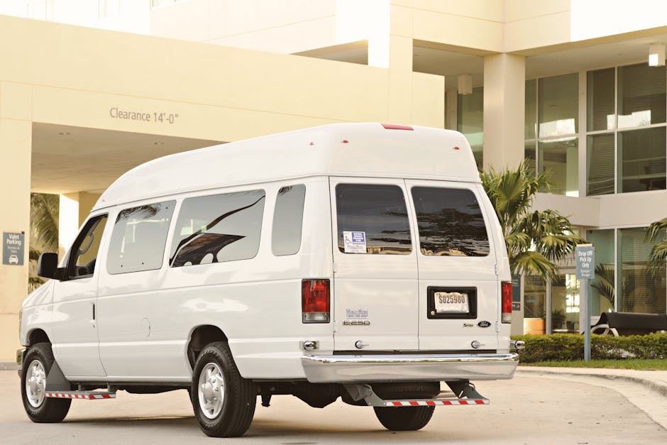 White passenger van parked in urban drop-off zone with palm trees and modern architecture.